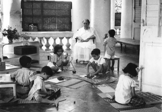 maria montessori dans Photographie en noir et blanc de Maria Montessori assise, observant plusieurs enfants indiens jouant et travaillant librement sur le sol d'une véranda, dans une Casa dei Bambini en Inde